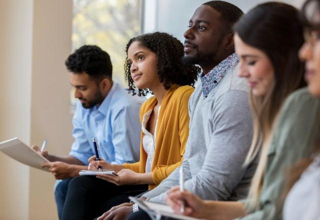 Four people sitting in a row listening to a speaker and taking notes