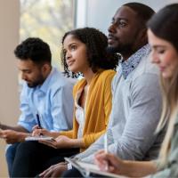 Four people sitting in a row listening to a speaker and taking notes