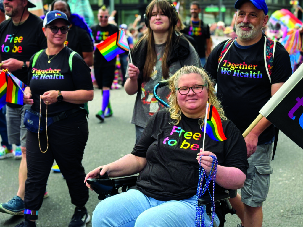 Group of people marching in Pride Parade wearing shirts that say Together We Health and Free to be in rainbow letters.