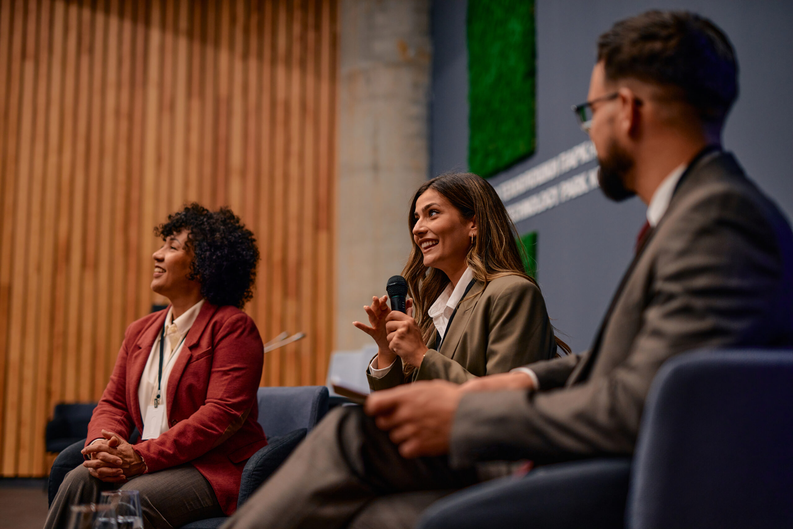 Three smiling people sitting in chairs speaking at a conference. Woman in middle holds a microphone.
